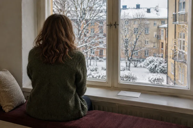 A person waiting by a window with an unopened envelope nearby.